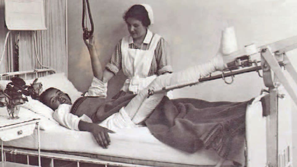 Eine alte Schwarzweiss-Aufnahme zeigt eine Krankenpflegerin am Bett einer Patientin, die einen Fuss eingegipst hat und sich von der Krankenpflegerin versorgen lässt.