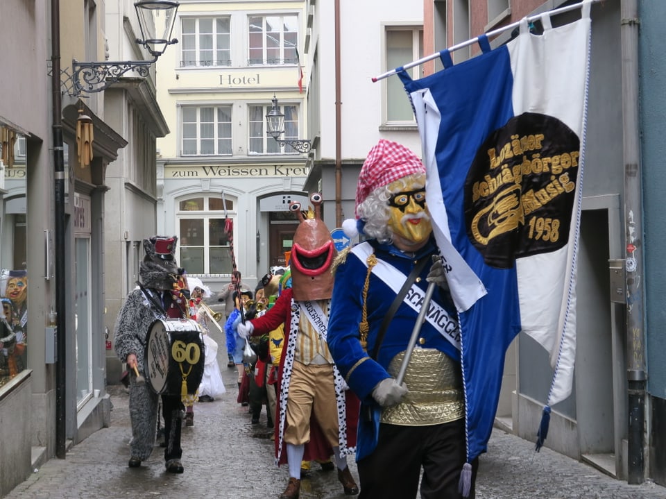Eine Luzerner Guggenmusig in einer Gasse mit einem Tambourmajor vorne, der eine Fahne hält. 
