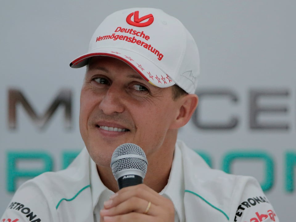 Michael Schumacher hält ein Mikrofon und lacht dabei.