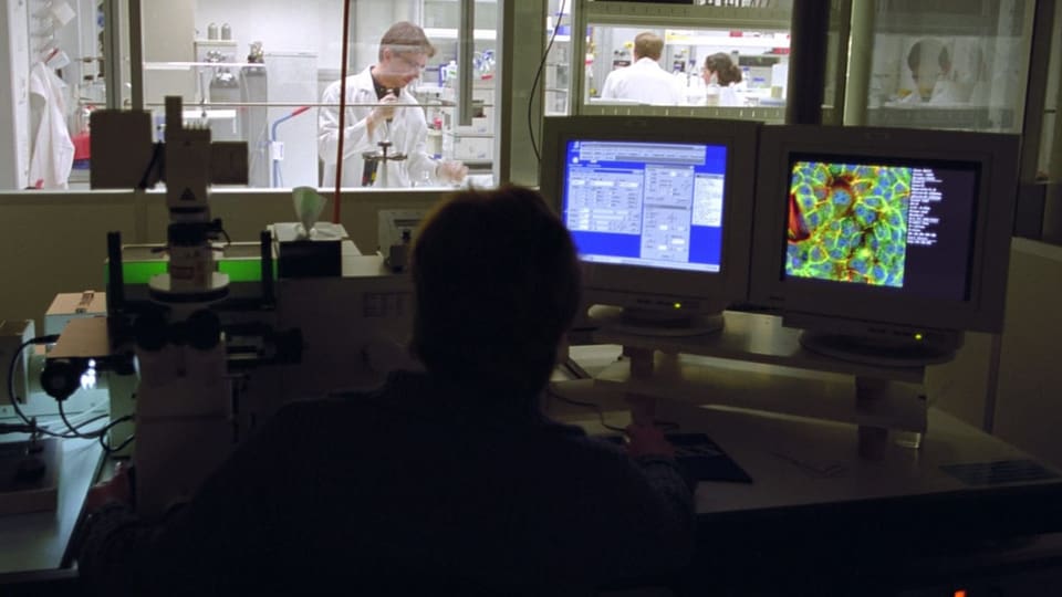 Ein Forscher arbeitet an einem Computer, hinter einer Glaswand arbeiten weitere Personen in einem Labor.