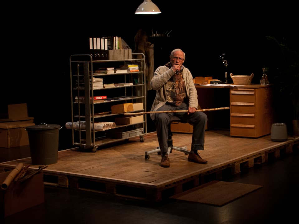 Martin Vosseler auf der Theaterbühne. Als Bühnenbild ist eine Bürosituation mit Schreibtisch 