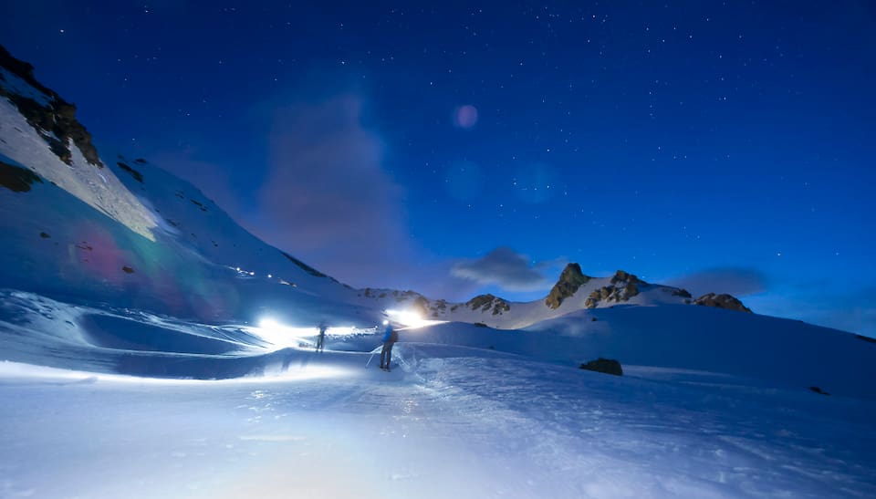 Nacht, Hochgebirge, viel Schnee, sternenklarer Himmel. Alles in schönen Blautönen. In der weite der Landschaft marschieren zwei Skitourenläufer. 