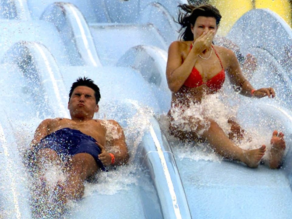 Ein Mann und eine Frau rutschen auf einer Wasserrutschbahn und das Wasser spritzt um ihre Ohren.