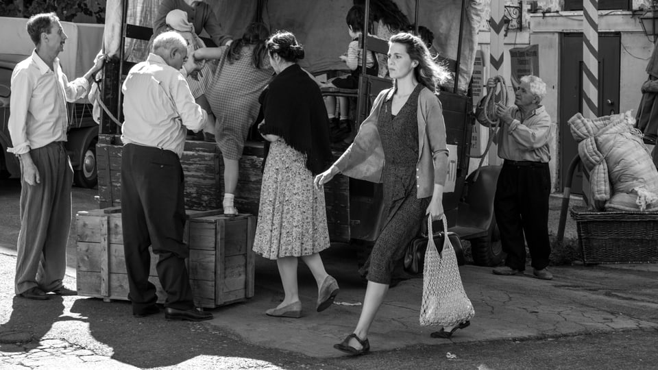 Eine Marktszene mit mehreren Menschen. Im Fokus eine junge Frau mit dunklem Kleid und Jacke, in der Hand eine Tasche.