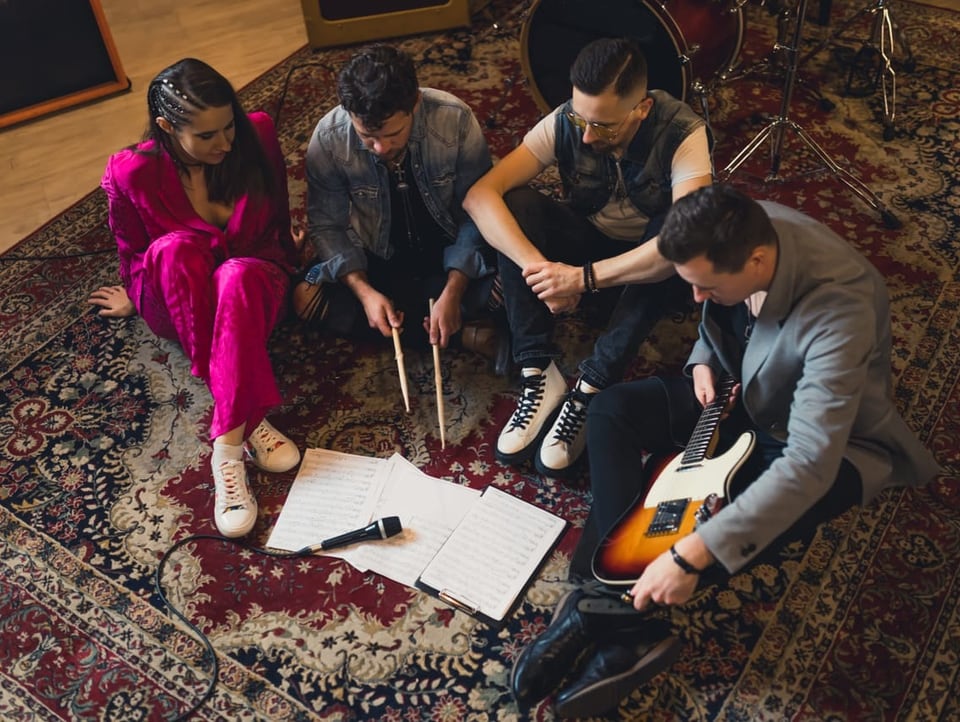 Bandmitglieder sitzen auf einem Teppich und sehen sich Notenblätter an. Vier Personen arbeiten an einem neuen Song