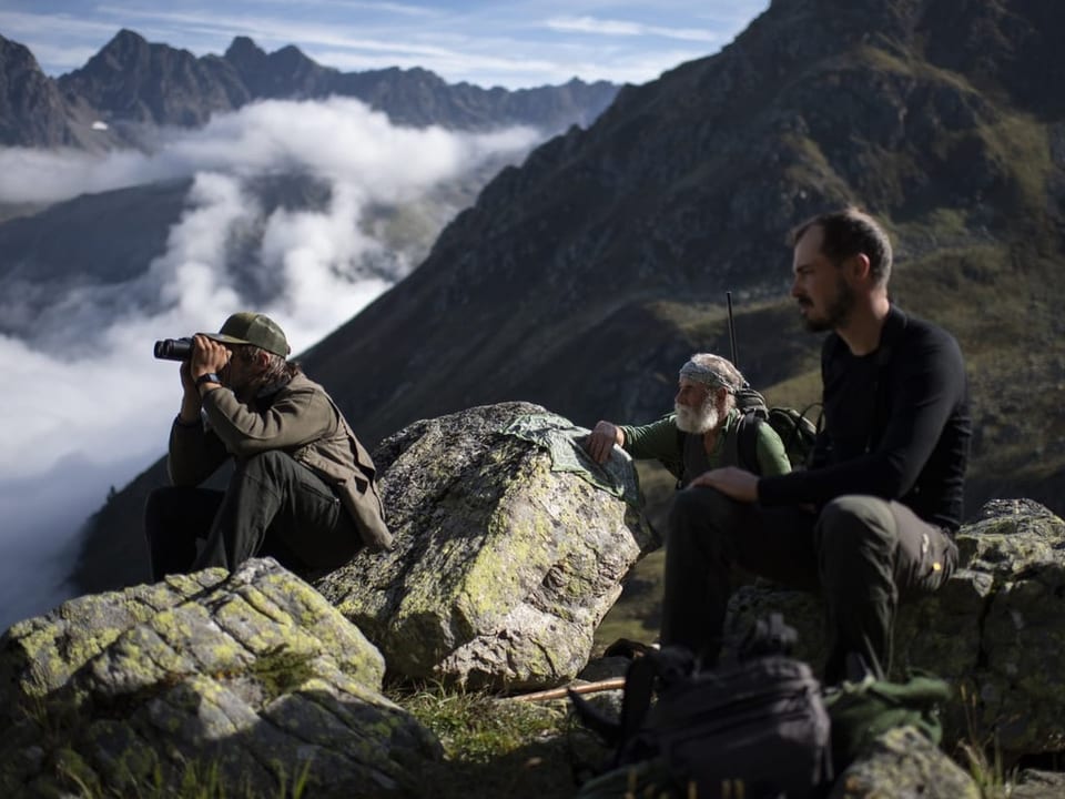 Drei Jäger sitzen in den Bündner Bergen auf der Pirsch, einer schaut durch ein Fernglas. 