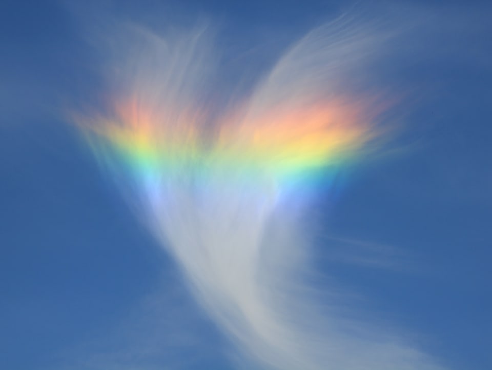 Eine Wolke, die einer Feder ähnelt, liegt am blauen Himmel. Ein farbiger Bogen zieht sich durch die Wolke.