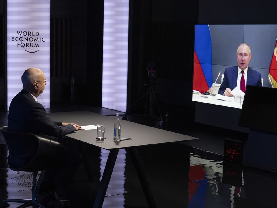 Klaus Schwab sitzt an einem Schreibtisch und blick auf einen Bildschirm, wo das Bild von Putin zu sehen ist.