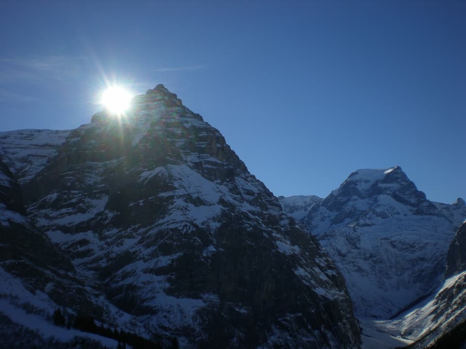 Der Himmel ist blau. Unten sieht man ein Bergmassiv mit etwas Schnee. Die Sonne ist knapp über einer Bergkante zu erkennen.