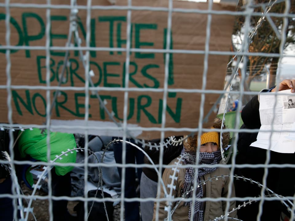 Hinter einem Drahtgeflecht und Nato-Stacheldraht sind Menschen, sie halten ein Kartonschild in die Höhe, auf dem steht  «Open the Borders - no return».