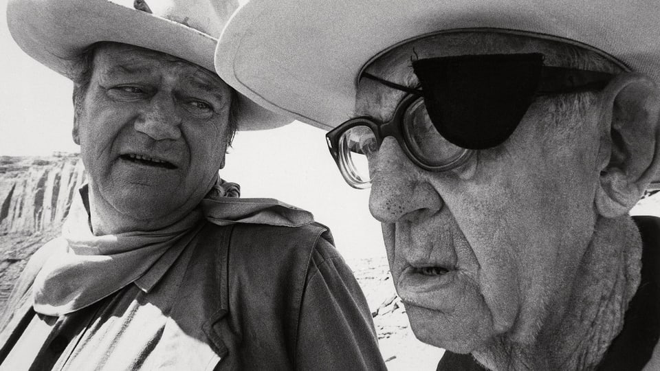 Ein Mann mit Cowboyhut spricht mit einem älteren Mann mit Brille und Augenklappe