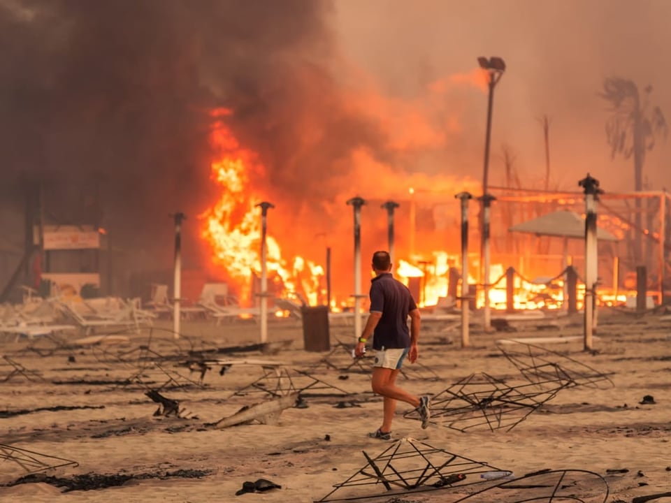 Ein Mann läuft am Strand. Im Hintergrund Flammen und verkohle Pfosten.
