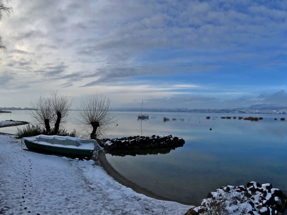 Spaziergang am frisch verschneiten Ufer vom Murtensee. Der Himmel zeigt ein paar Wolkenlücken. Der See ist spiegelglatt.