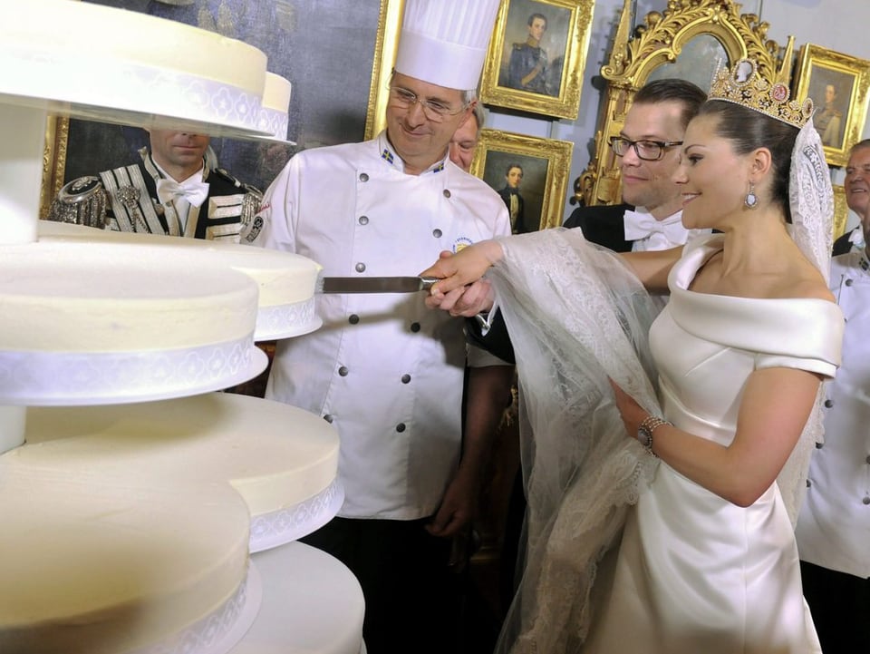 Victoria und Prinz Daniel schneiden gemeinsam ein Messer halten einen Kuchen an. Im Hintergrund stehen Köche. 
