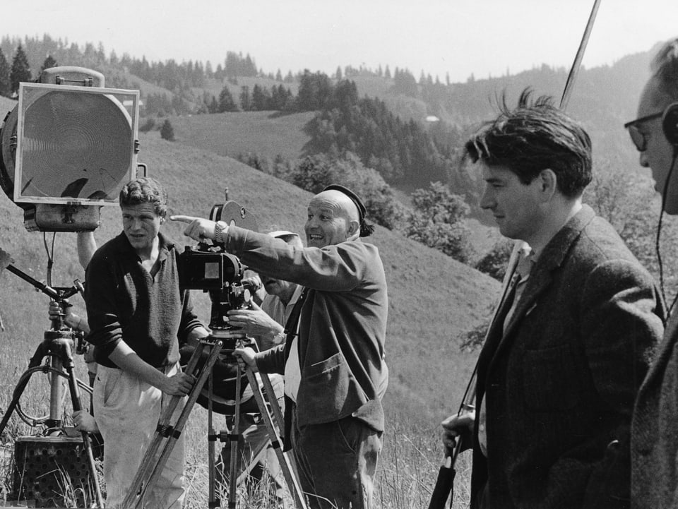 Franz Schnyder zeigt neben der Kamera stehend mit energischem Blick auf das Set. Der Kameraassistent sowie ein Filmscheinwerfer stehen neben der Kamera. Im Vordergrund der Tonmeister.