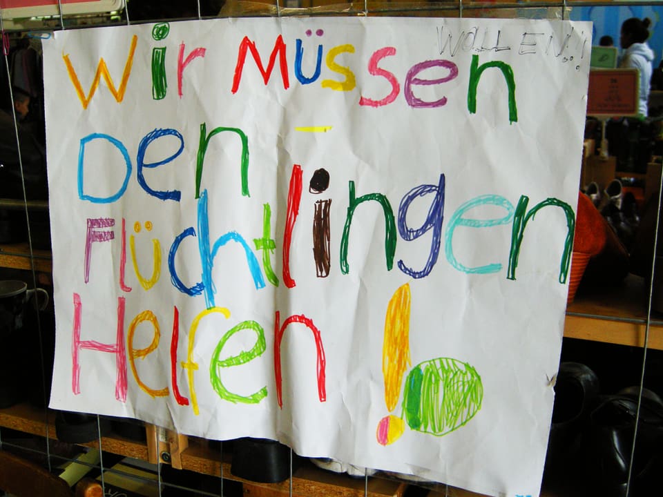 Plakat in Kinderschrift: Wir müssen, nein wollen!, den Flüchtlingen helfen.