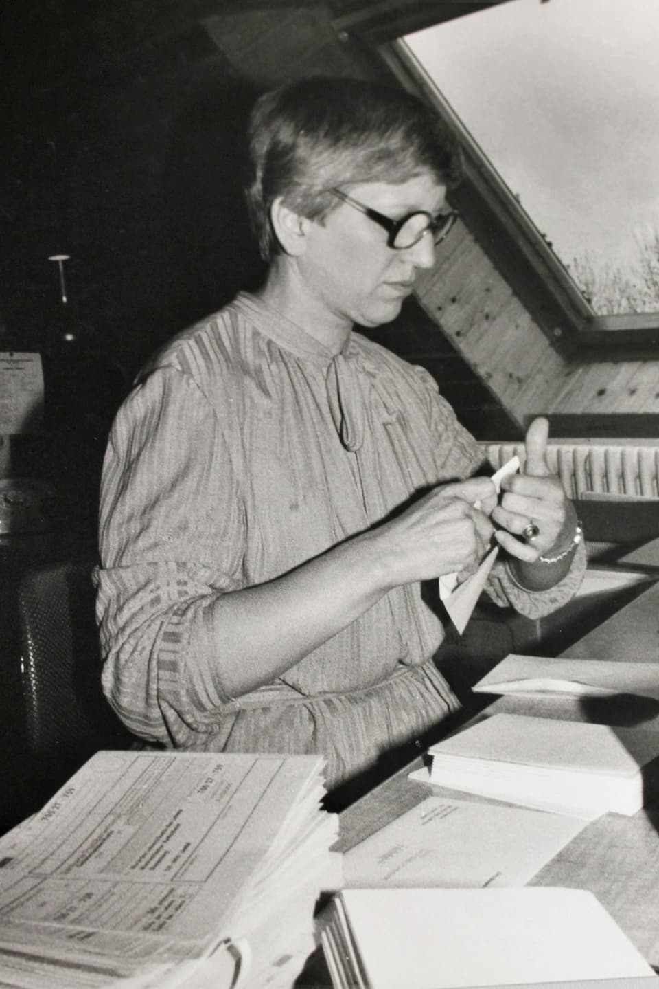 Schwarzweiss Bild: Frau mit Kleid, kurzen Haaren und grosser Brille sitzt an einem Schreibtisch und tütet gerade einen Brief in ein Couvert ein.
