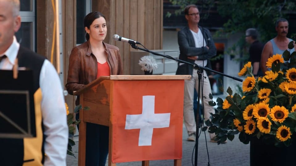Eine junge Frau hinter einem Rednerpult, das eine Schweizer Flagge ziert.