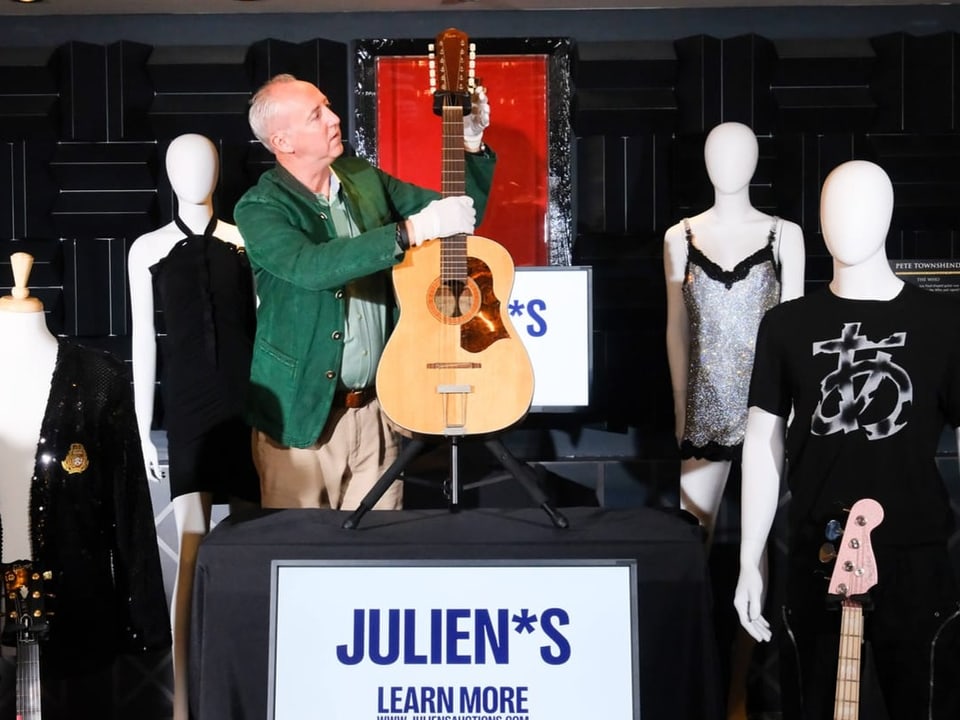 Mann präsentiert Gitarre neben Mannequins mit Kleidern auf Auktion.