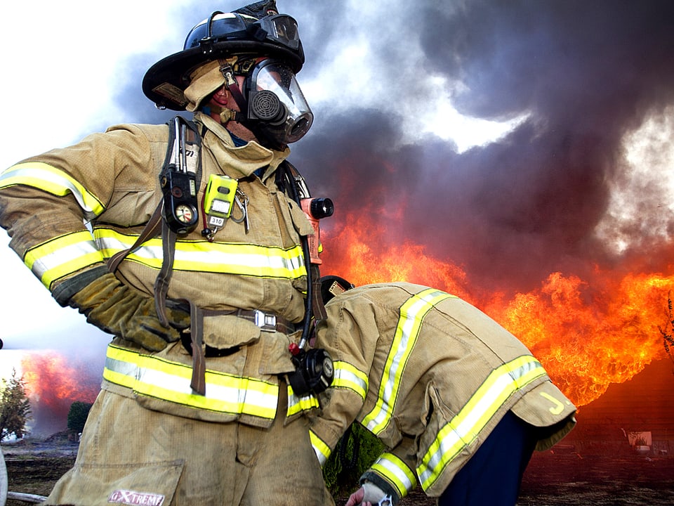 Zwei Feuerwehrmänner in voller Montur mit Schutzmasken. Im Hintergrund tobt die Feuersbrunst.
