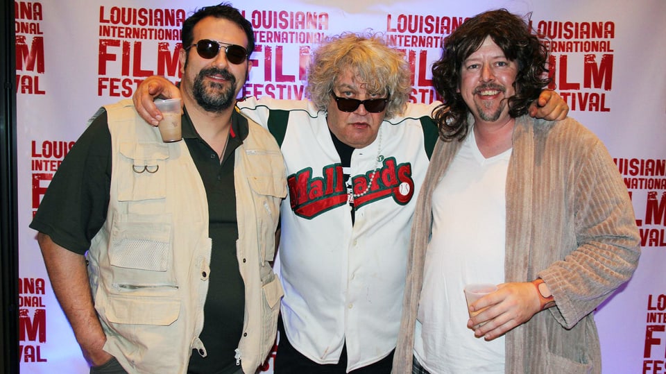 Drei Männer posieren für das Foto in einer Reihe. Derjenige rechts und derjenige links sind als zwei Charaktere aus dem Film verkleidet, in der Mitte steht Jwff Dowd mit Hemd und Sonnenbrille.