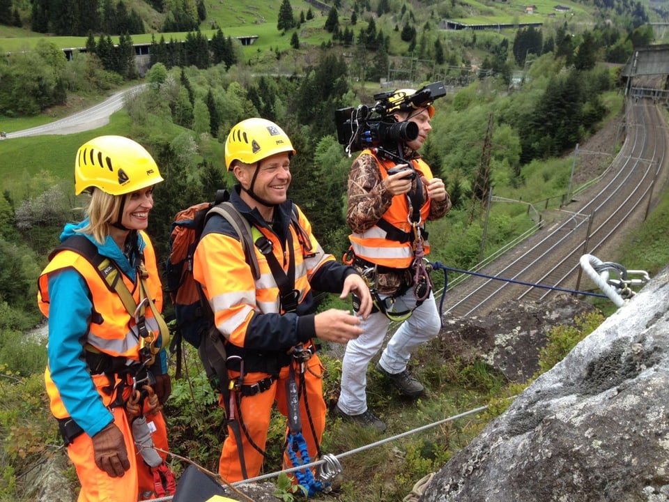 Drei Leute hängen im Fels, darunter eine Eisenbahnlinie. Ein Mann trägt eine Kamera auf den Schultern.