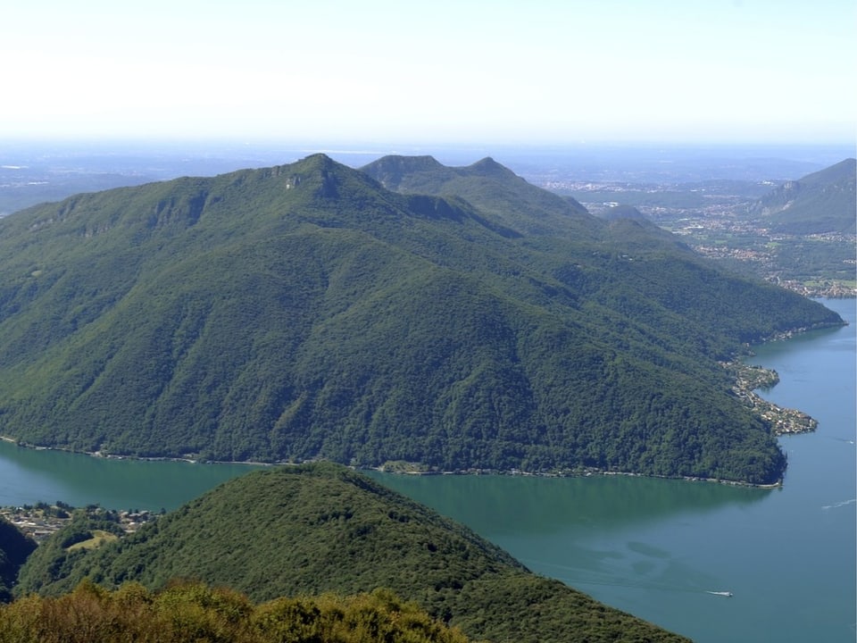 Blick auf einen grossen bewaldeten Hügel im Kanton Tessin. Darum liegt ein See.