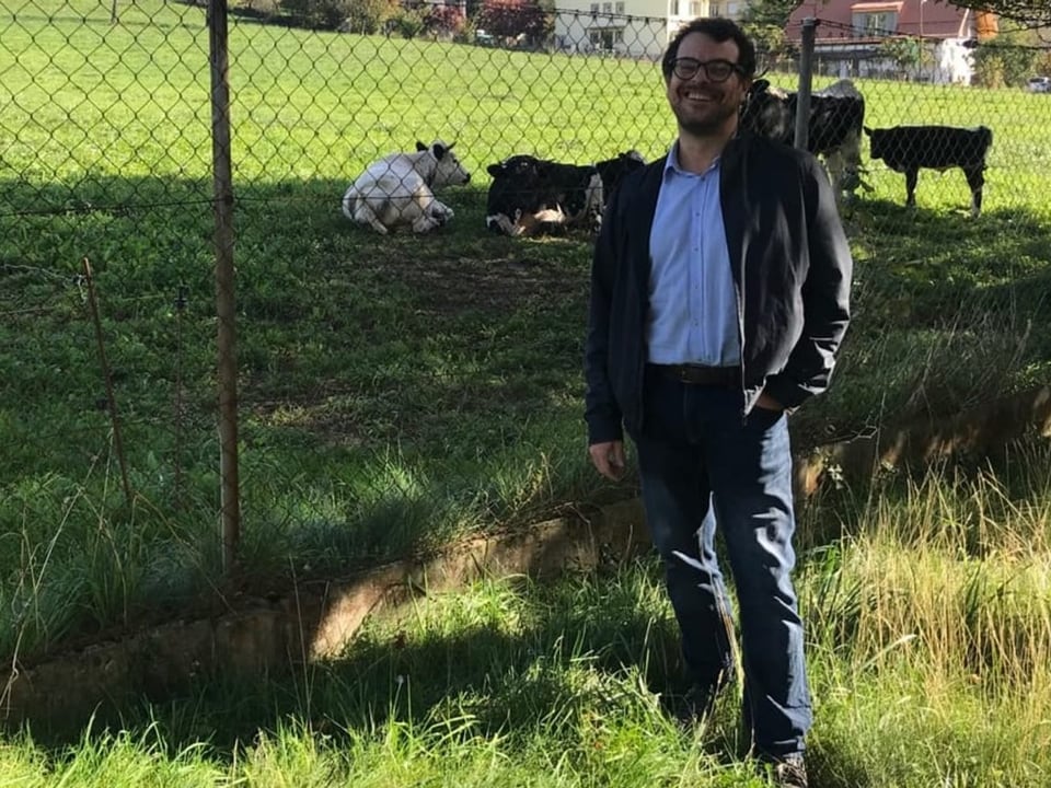 Mann mit brauner Jacke und Jeanshosen steht vor einem Zaun auf einer Wiese. Im Hintergrund sind Kühe.
