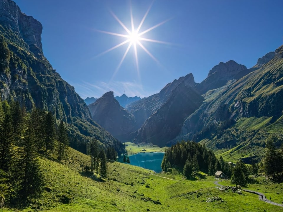 Bergsee in grünen Wiesen und felsigen Bergen. Die Sonne scheint am wolkenlosen Himmel. 