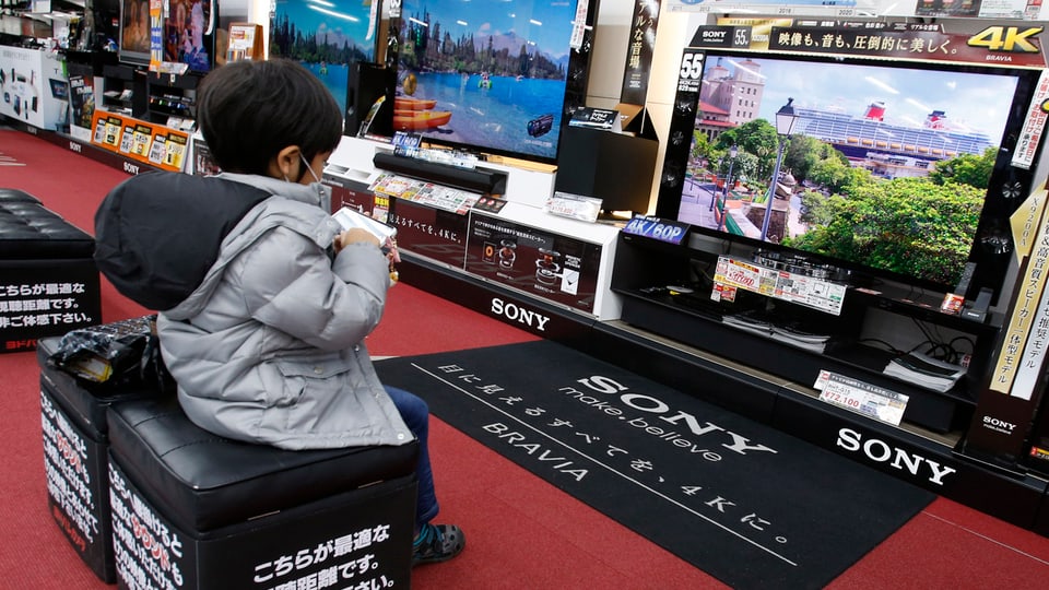 Ein kleiner Junge spielt auf seinem Smartphone vor einem grossen TV-Bildschirm.