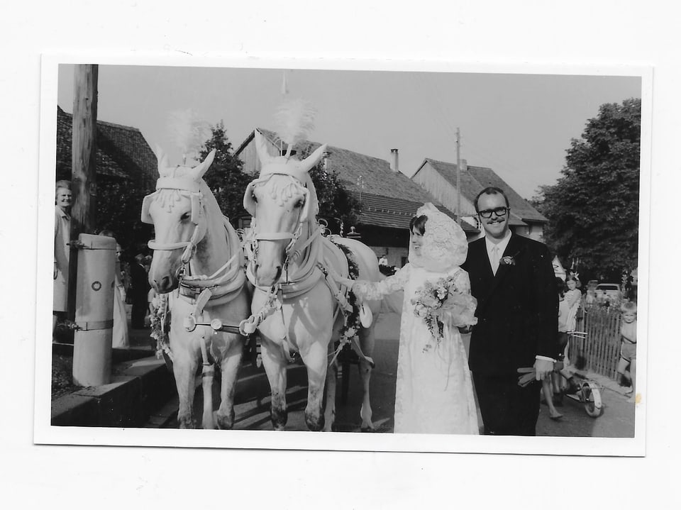 Auf einem Schwarz/Weiss-Foto steht das Brautpaar neben zwei weissen Pferden, welche voreine Kutsche gespannt sind.