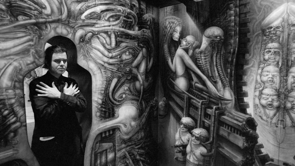 Breaking Boundaries Maison d'Ailleurs Hosts First Tattoo Convention in a Swiss Museum 6 Ein Mann Alters überkreuzt beide Arme vor seiner Brust. Er steht in einem Zimmer, das mit Gruselbildern ausgemalt ist.