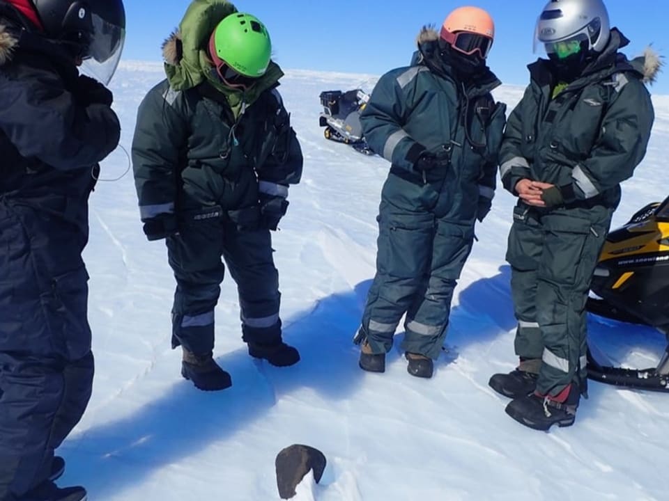 Menschen in dicken Thermoanzügen stehen im Halbkreis um einen Meteoriten im Schnee