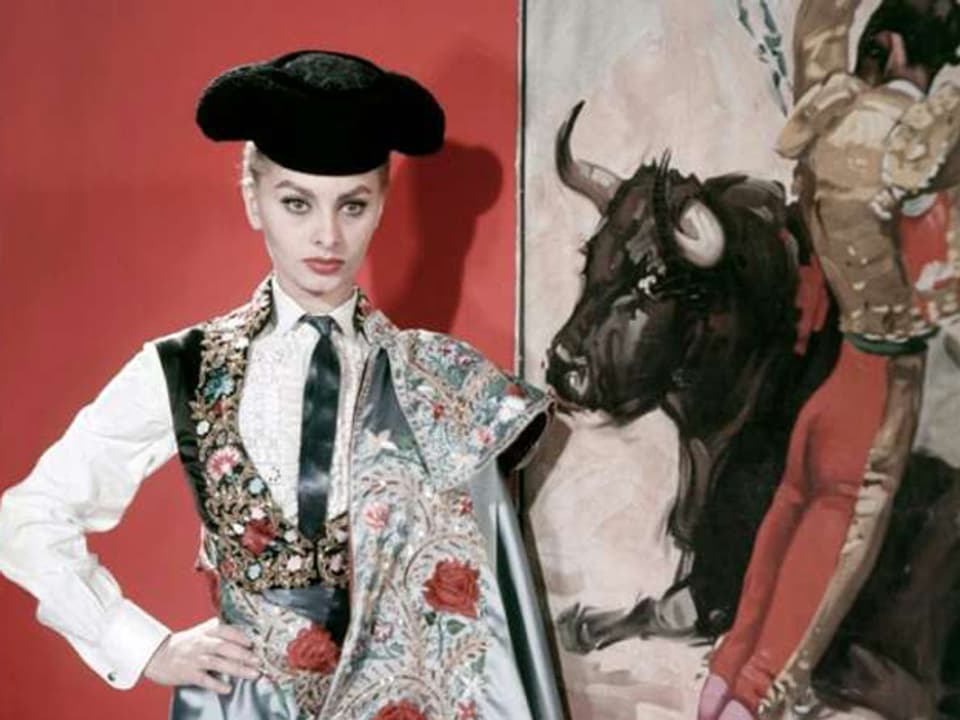 Eine geschminkte Frau steht in Torero-Kostüm vor einem Bild, auf dem ein Stier zu sehen ist.