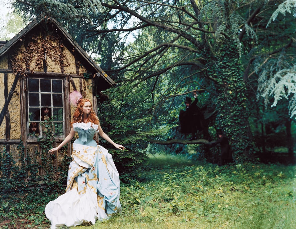 Eine Frau mit langen roten Haaren trägt ein blau, weisses und goldiges Kleid. Sie läuft über grünes Gras und an einer märchenhaften Hütte vorbei.