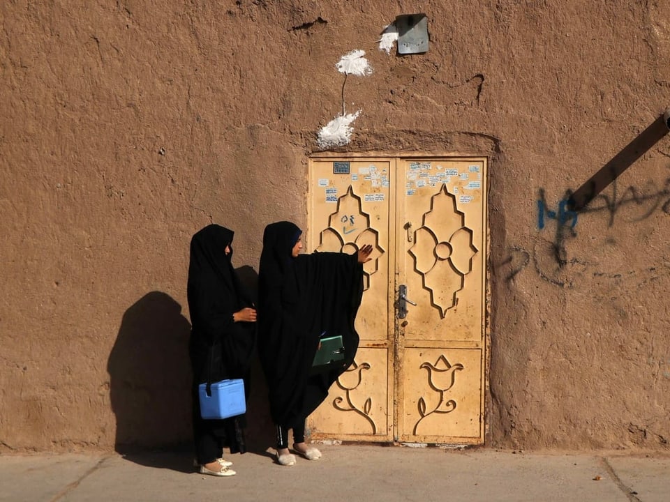 Zwei Frauen tragen eine blaue Kühlbox (vermutlich mit einer Polioimpfung darin) zu einer Haustüre. Beide Frauen tragen eine Burka.