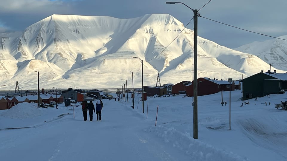 vorne verschneite Strassen, hinten einige Häuser mit verschneiten Dächern, dahinter weisse Berge. Drei Menschen laufen.