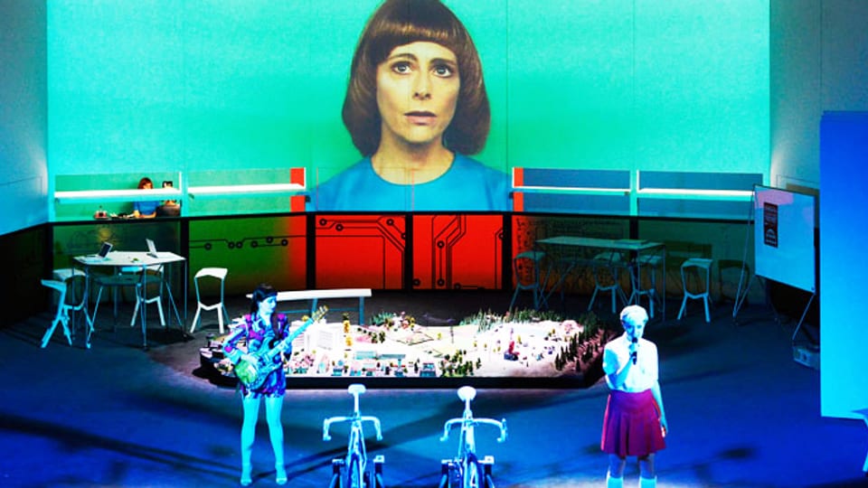 Zwei Fahrräder stehen am Bühnenrand daneben eine junge Frau. Im Hintergrund ist das Bild einer andern Frau gross an die Wand projiziert. 