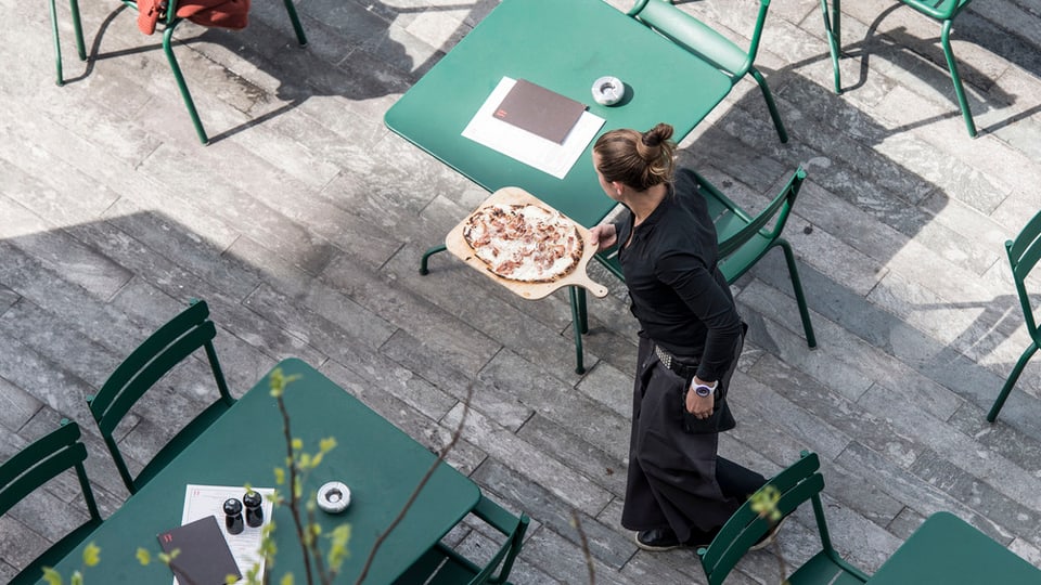 Kellnerin serviert eine Pizza auf einer Gartenterrasse mit vielen unbesetzten Plätzen.
