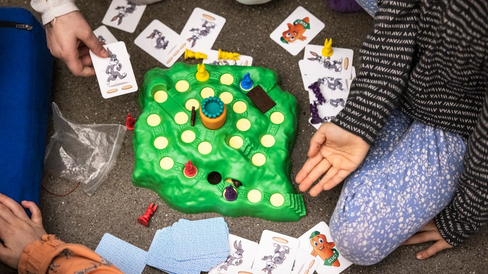 Ansicht von oben, ein Spiel mit grüner Plastikinsel und Spielkarten. Rund herum sitzen Kinder, Beine und Hände sichtbar.