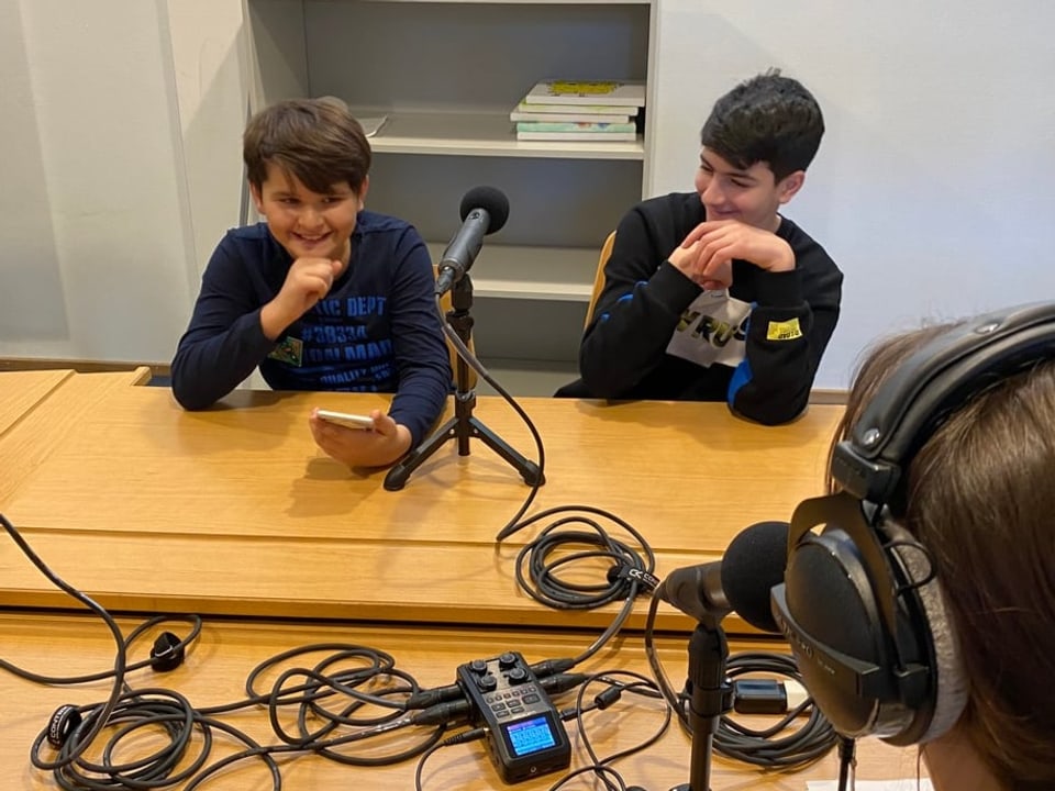 Zwei Jungen, beide dunkel gekleidet, sitzen lachend vor einem Mikrofon. Der Junge links hält sein Handy in der Hand.