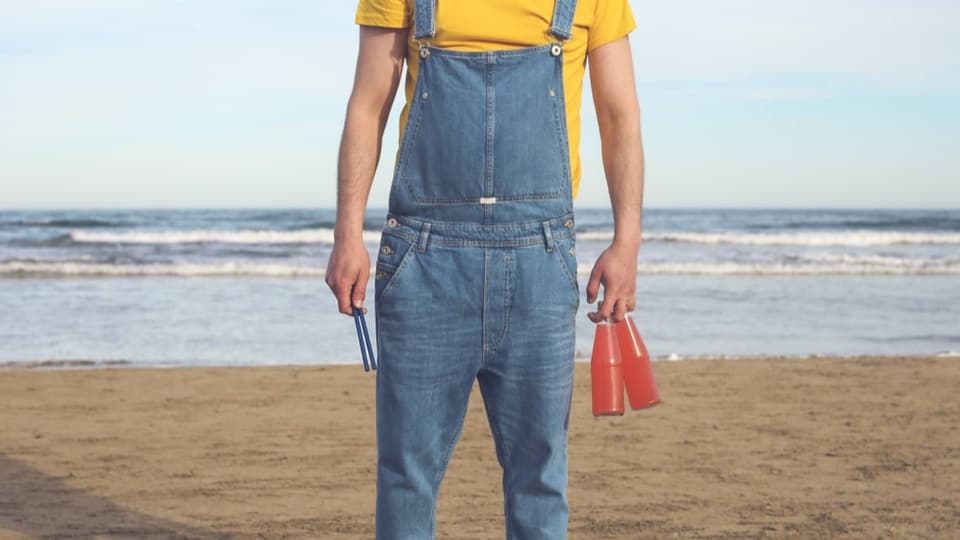 Mann mit Jeans-Latzhose steht am Strand, dreht dem Meer den Rücken.