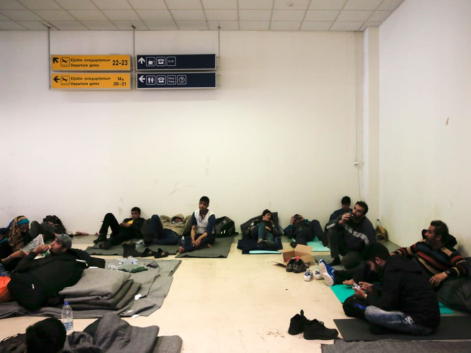 Flüchtlinge sitzen am Boden auf dünnen Matten in einer Halle des alten Athener Flughafens. 