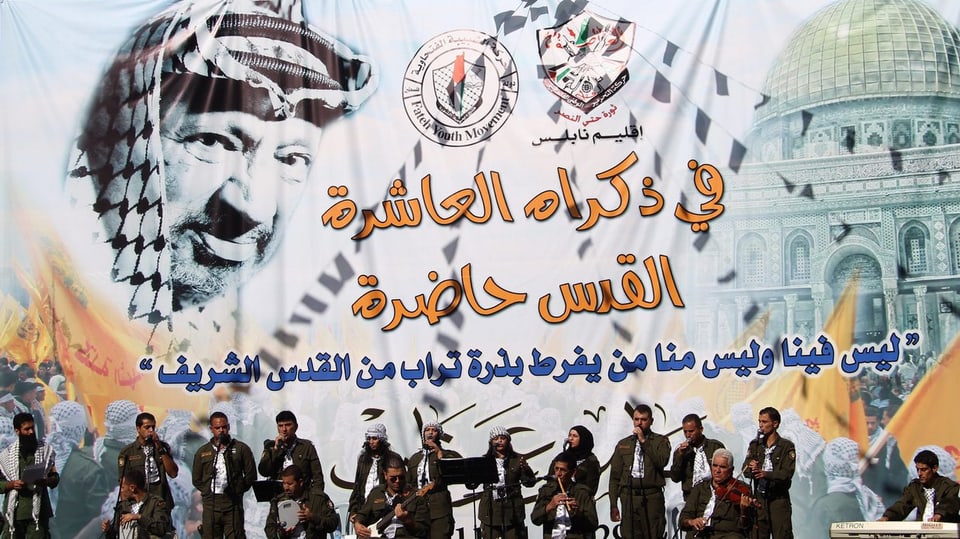 Eine Musik-Band aus mehreren Mitgliedern bestehend spielt vor dem Hintergrund eines Arafat-Porträts