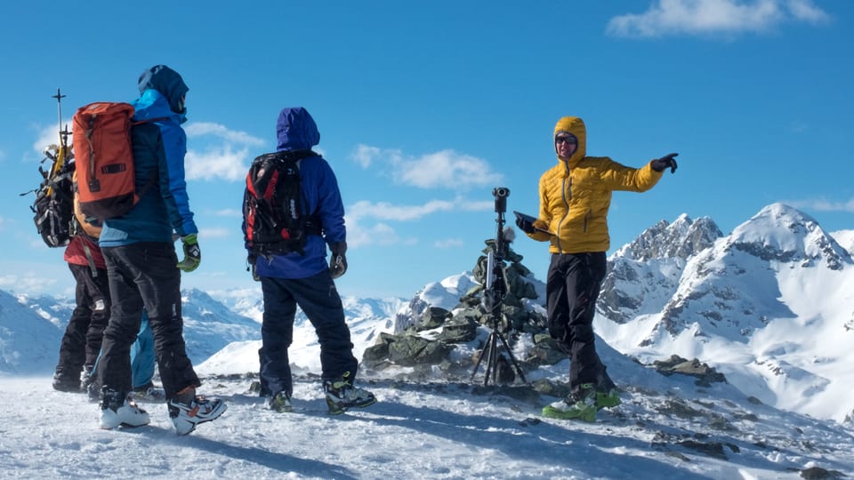 Eine Gruppe von Menschen steht auf einem Berggipfel im Winter. Im Hintergrund sind weitere Gipfel der Alpen sichtbar. In der Mitte des Bildes steht eine Kamera auf einem Stativ. 