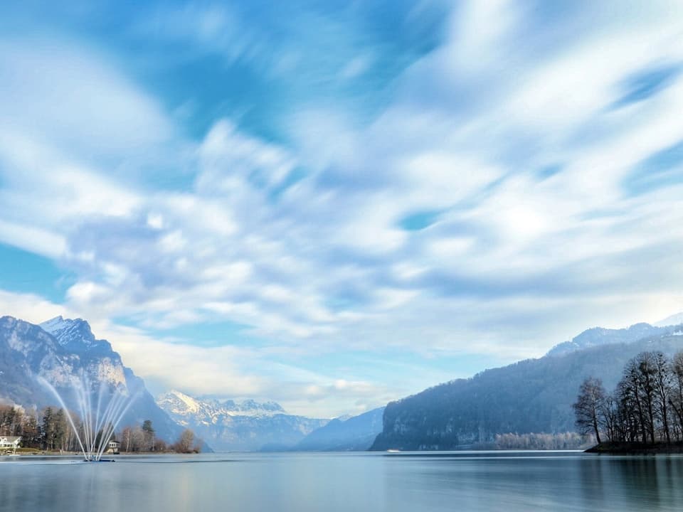 Blick auf den Walensee mit den Bergen im Hintergrund. Links im Bild sieht man noch einen Springbrunnen im See, rechts am Ufer sieht man dunkle Bäume. Der Himmel ist ein Mix zwischen Blau und Wolken.