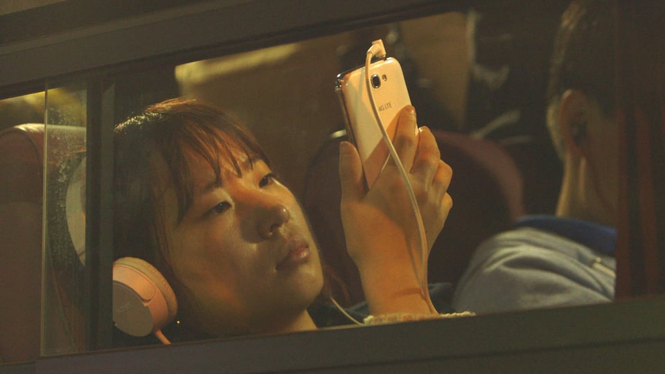 Eine junge Asiatin mit Kopfhörer in einem Bus, den Blick hat sie auf ihr Smartphone gerichtet.
