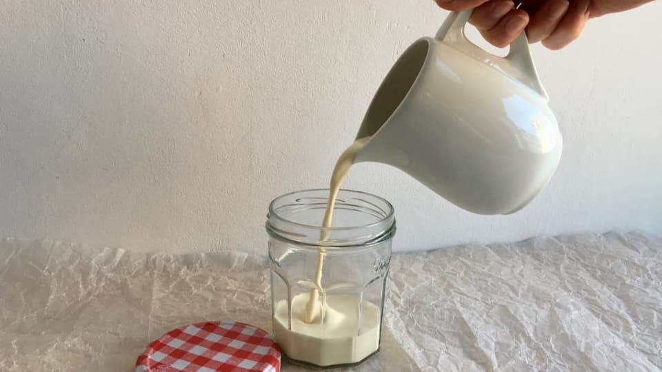 Jemand giesst aus einem kleinen Krug Rahm in eine Konfitüren-Glas