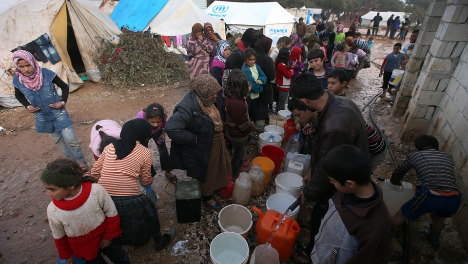 Syrische Flüchtlinge stehen in einem Flüchtlingslager um zahlreiche Behälter.