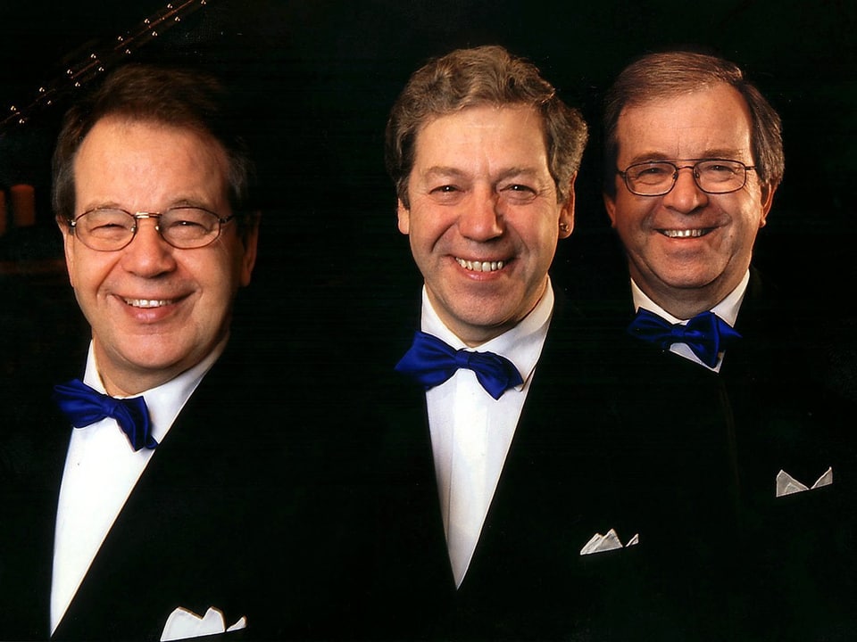 Drei Männer in dunklen Anzügen, weissen Hemden und blauer Fliege.